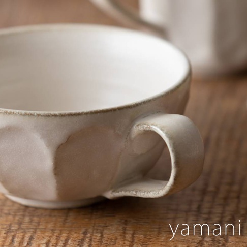 一個帶有六邊形紋理的米白色陶瓷湯杯，帶有粗糙的邊緣和一個彎曲的把手，放在木製桌面上。