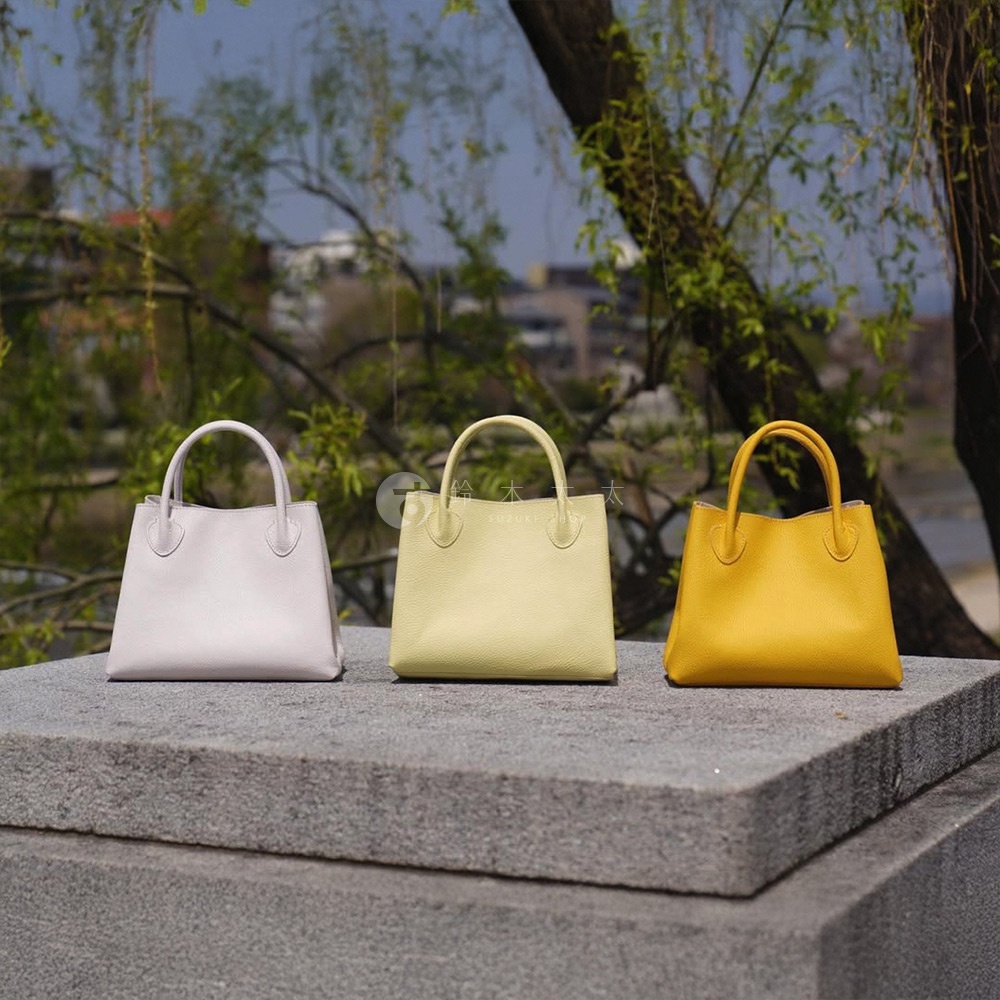 三個皮革手提托特包，分別為淡紫色、淺黃色和亮黃色，擺放在灰色石台上，背景為綠葉與樹木。