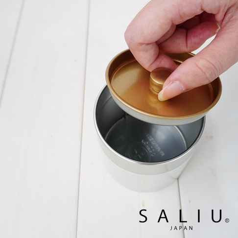 銀色錫製茶葉罐，附金色圓形掀蓋，罐身外部為霧面銀色，內部為銀色，蓋子內側為金色，由手部拿起蓋子準備蓋上。