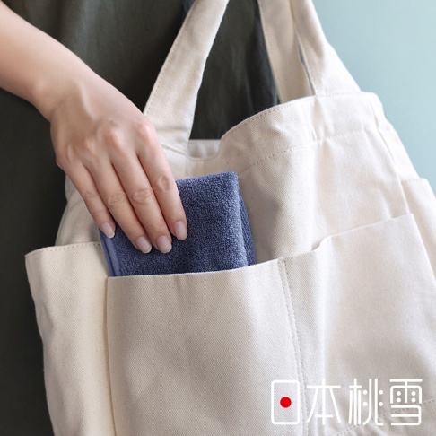 一個人的手正在將一條藍色毛巾放入米白色帆布托特包的外部口袋，包的右下方印有「日本桃雪」的字樣。