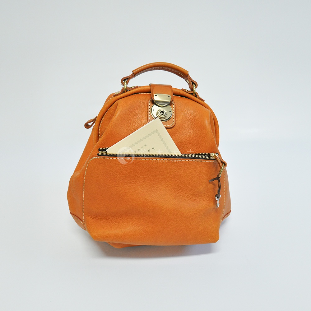職人手作皮革輕旅行後背包 R-144，橘棕色，皮革材質，前方有一個拉鍊口袋，內有一個小卡片，上方有提把和金屬鎖扣，側邊掛有小鑰匙吊飾。