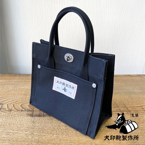 黑色純棉帆布迷你托特包，頂部有提把和旋轉鎖扣，正面有一個帶有品牌標籤的翻蓋口袋，口袋上裝飾有銀色鉚釘。