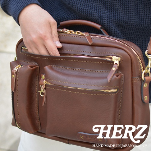 職人手作皮革兩用多功能收納單肩包，採用咖啡色皮革，設有頂部提把、多個拉鍊隔層和前置口袋，搭配金色拉鍊和五金配件，展現精緻工藝。