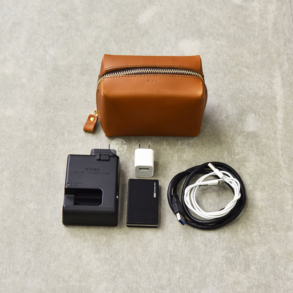 一個淺棕色的皮革收納包，旁邊擺放著一個黑色 Nikon 電池充電器、一個白色充電頭、一個黑色行動硬碟和兩條充電線。