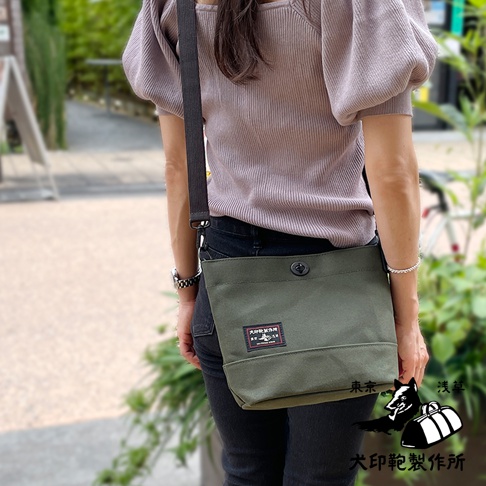 一位女性斜挎著一個軍綠色純棉帆布迷你側背包，包上有一個黑色鈕扣和一個印有日文文字及狗圖案的標籤。