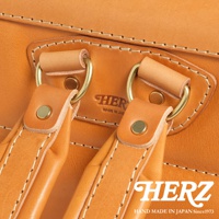 HERZ 品牌標誌壓印在淺棕色皮革上，下方有 HAND MADE IN JAPAN Since1973 字樣，圖片展示了包款的提把連接處，使用黃銅金屬環和鉚釘，縫線細緻。