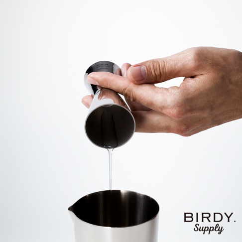 一只手正握著一個不鏽鋼雙頭刻度量酒器，將液體從較小的量杯傾倒到較大的量杯中。量酒器表面光滑，有品牌標誌。