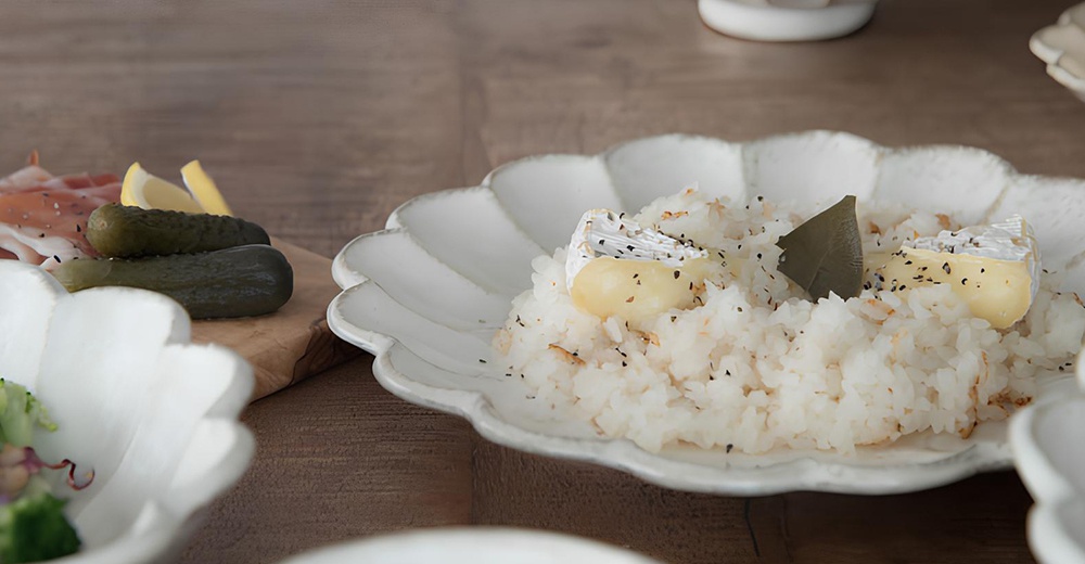 白色陶瓷主食盤，帶有六瓣花型邊緣，盤中盛有白飯、起司和裝飾葉片，盤子質感樸實。