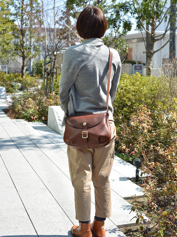 棕色皮革馬鞍包，附有金色扣環和可調式肩帶，由一位身穿灰色毛衣和卡其色褲子的女性斜挎著。