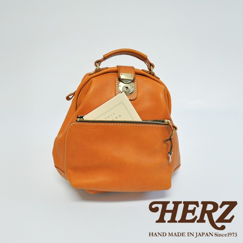 橙色皮革手提後背包，具有頂部提把、金色金屬鎖扣及拉鍊前袋，袋中插有一本淺色書籍。