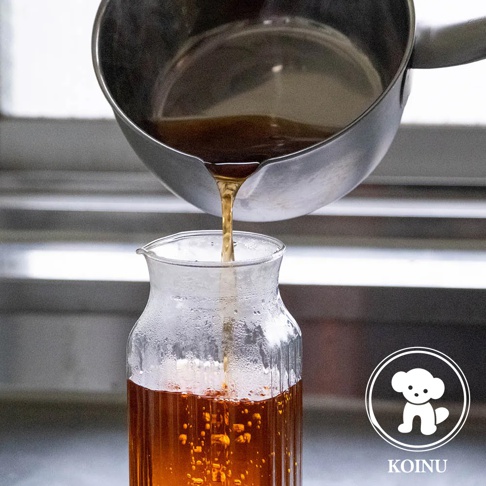 一個不鏽鋼雪平鍋傾倒著琥珀色液體，流入一個帶有細緻紋路的玻璃瓶中。玻璃瓶內裝有相同顏色的液體，瓶身佈滿細小的水珠。