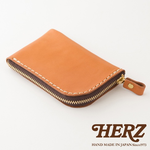 這是一款日本HERZ手工製作的皮革拉鍊零錢包，呈現棕色皮革質感，包身採用手工縫線設計，側邊配有同色皮革拉環，下方標示著HERZ品牌字樣與產地資訊。