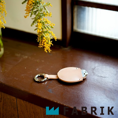 FABRIK 皮革圓型收納鑰匙圈，附有銀色掛環，放在深棕色木質檯面上，背景有黃色花束和窗戶