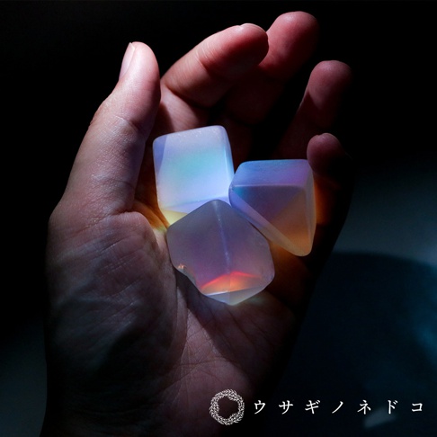 手掌中握著三個透明、多面體的合光稜鏡石，呈現漸層的藍、粉、橘色光暈，材質光滑，邊緣清晰。