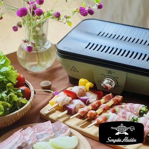 輕便式迷你烤盤，淺綠色機身搭配黑色烤網，設有火力調節旋鈕。旁邊擺放著各式烤串、生肉片、蔬菜和一束紫色小花，營造出居家燒烤的氛圍。