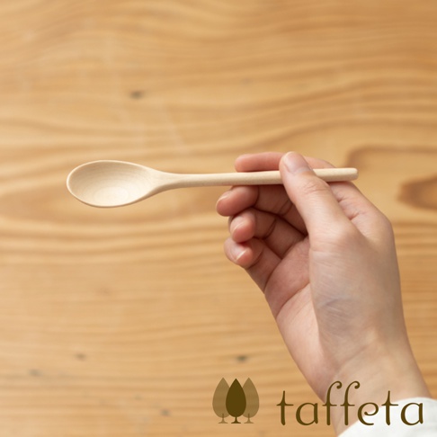 一隻手拿著一支淺色楓木茶匙，茶匙勺部圓潤，木紋清晰，手持部分細長，背景為淺木紋木板。