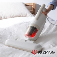 白色IRIS OHYAMA除塵蹣機，搭配透明紅色集塵筒，可見內部濾網。機身設有操作按鈕，底部連接寬扁吸頭，放置於白色床鋪上，背景為木質床頭。