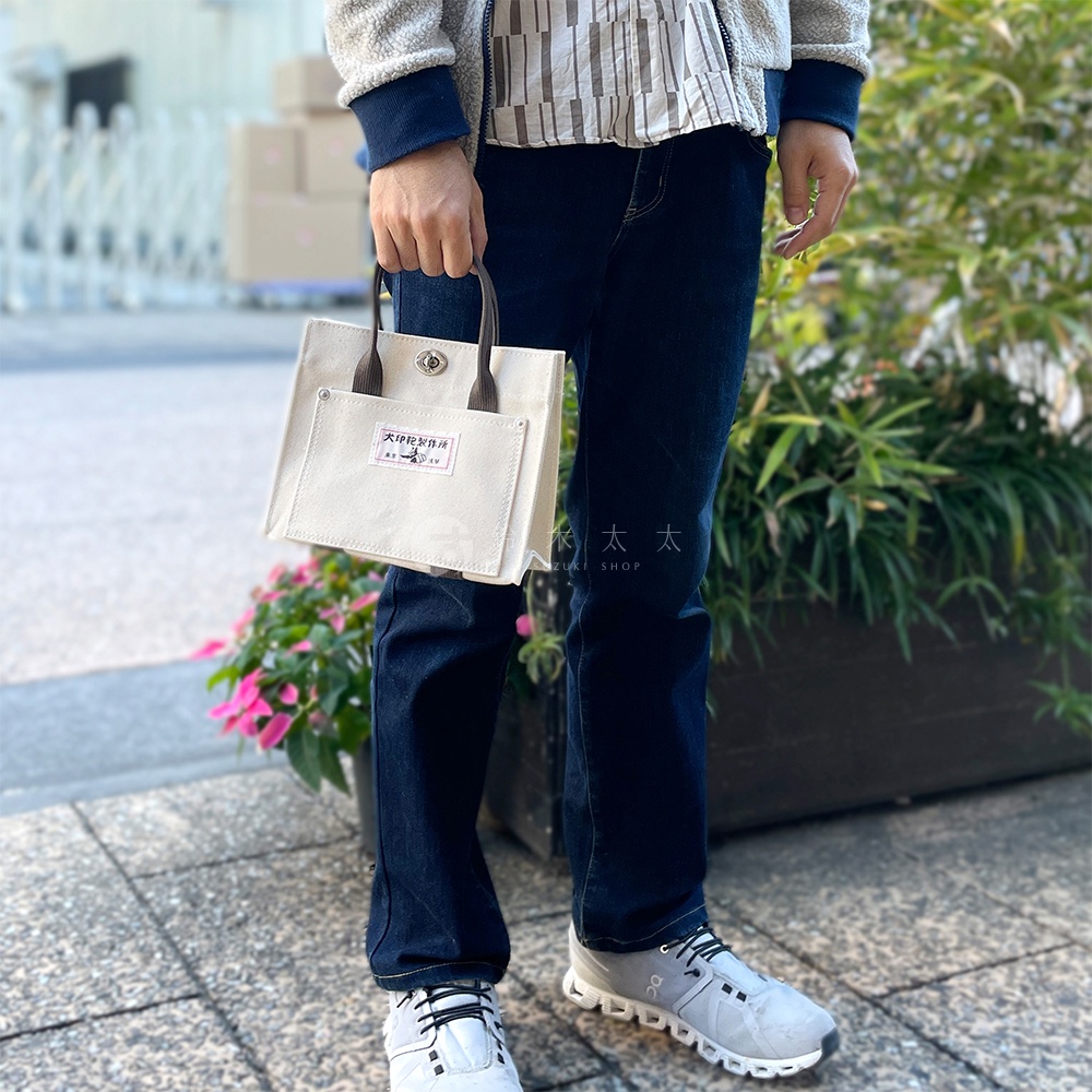 一人手持一個米白色純棉帆布迷你托特包，包包有一個深棕色提把，正面有一個同色系小口袋，口袋上方有一個標籤，標籤上印有日文文字。