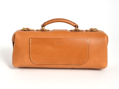 職人手作皮革橫長形單肩包，呈現溫潤的淺棕色皮革，包身設計簡潔，頂部配有手柄，兩側有金屬扣環，正面有一個方形外袋，上面壓印有品牌標誌。