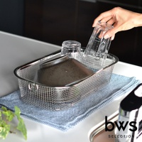 簡約風格的長方形不鏽鋼瀝水籃，內有兩個透明玻璃杯和一個深色碗，瀝水籃置於藍色紋理的布墊上，一只手正將一個玻璃杯放入籃中。