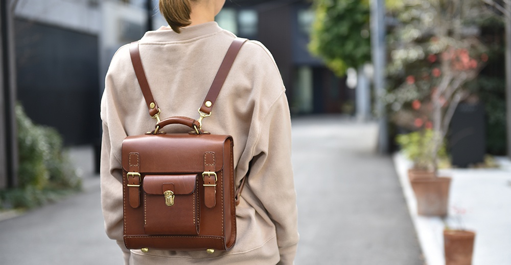 皮革學院風迷你後背包，使用棕色皮革製作，有單一翻蓋和金屬鎖扣，兩側配有可調節式皮質肩帶，可背在肩上。