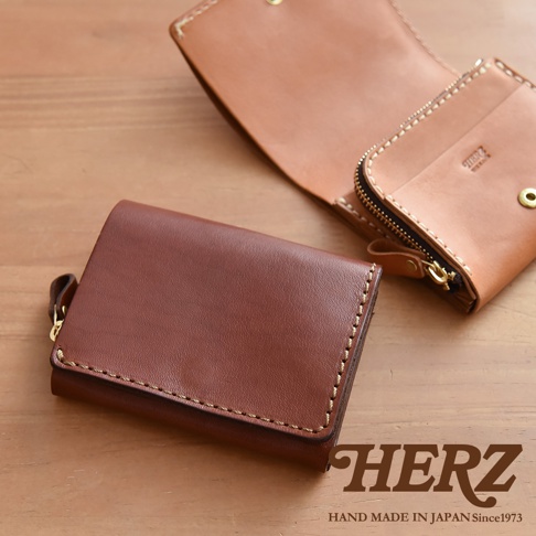 深棕色職人手作皮革小型雙折短夾，配有同色系車縫線及金色拉鍊，另一款淺棕色皮夾展開，露出內部卡槽與品牌壓印。