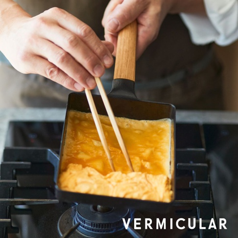 黑色琺瑯鑄鐵玉子燒鍋，配有木製手柄，正用於烹煮蛋液，鍋內蛋液呈現金黃色，並有氣泡。