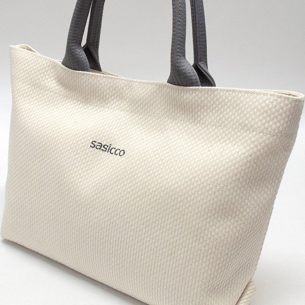 米白色三河木綿刺子織托特包，配有深灰色織物提把，包身有黑色「sasicco」字樣。