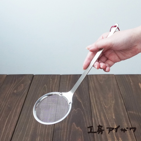 一只手正持著一個銀色不鏽鋼萬用撈油勺，勺頭為橢圓形，內嵌細密網篩，勺柄末端有一個紅色掛環。