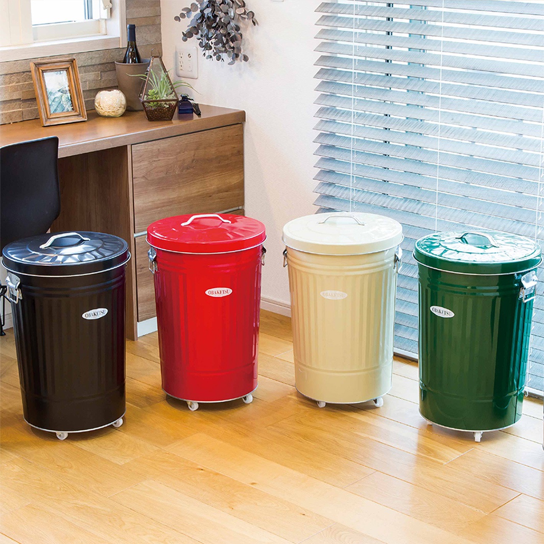 四個不同顏色的OBAKETSU手工製附輪垃圾桶，擺放在木質地板上，背景是百葉窗和辦公桌。