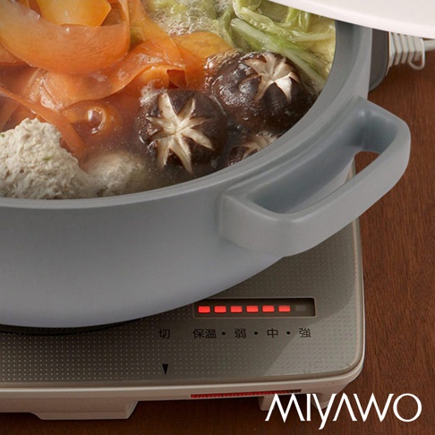 灰白色陶土湯鍋，鍋內有香菇、丸子、紅蘿蔔片和青菜，湯鍋置於有加熱指示燈的爐具上。