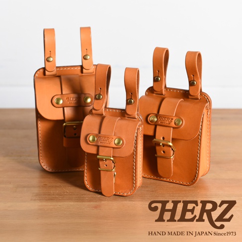 職人手作皮革經典腰袋 E-49，由鞣製皮革製成，呈現自然棕色。款式小巧，帶有翻蓋和金屬扣環，並有兩個皮革掛袢，可繫於腰帶上。共有三種尺寸，圖片中展示了不同尺寸的腰袋。