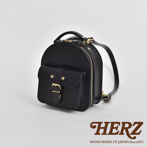 黑色皮革迷你後背包，圓潤造型，正面有翻蓋式口袋，配有黃銅釦環及皮帶。側邊有拉鍊開合，雙肩背帶可調。