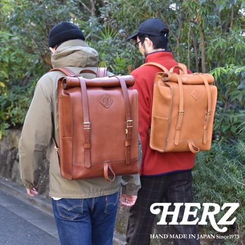 兩款職人手作皮革後背包，分別為深棕色與淺棕色，皆採用捲口設計，配有皮質揹帶、金屬釦具及側袋，適合戶外活動使用。