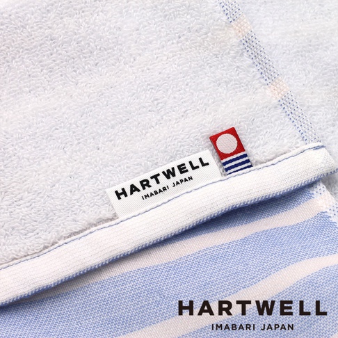 一張特寫視角的照片，展示了帶有 Hartwell Imabari Japan 標籤的純棉紗布浴巾，布料呈現白色與淡藍色的條紋質感，並附有一枚印有紅圓圈與藍色線條的織標，右下角亦印有該品牌名稱與產地標示。
