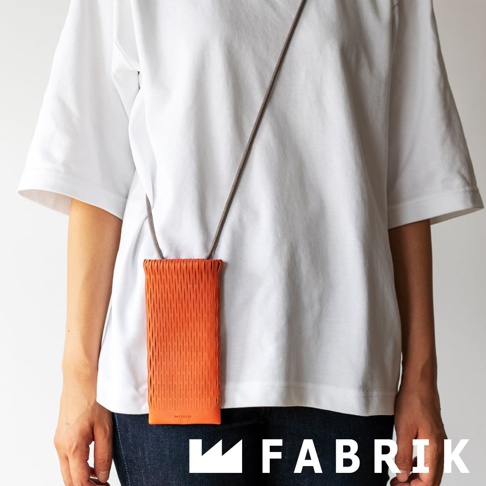 FABRIK 品牌標誌和由細條紋編織成的橘色皮革手機袋，由一位身穿白色 T 恤和牛仔褲的人配戴。