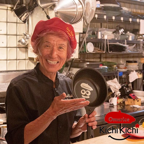 一位廚師手持一個黑色日式煎鍋，鍋底印有「Omurice」字樣和可愛的卡通圖案。鍋身為黑色，材質疑似不沾塗層。