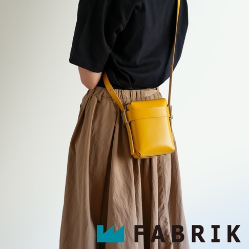 FABRIK 品牌字樣旁，一人身著黑色上衣與棕色長裙，斜挎著一個黃色皮革迷你斜背包。