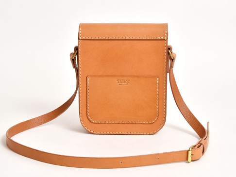 淺棕色皮革翻蓋迷你單肩包，正面有一個貼袋，袋蓋上有品牌標誌，包身線條簡潔，配有可調節長度的皮革肩帶。