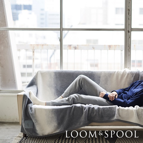 一件淺灰色與白色漸層的抗菌短絨舒柔單人毯，覆蓋在沙發上，一人穿著灰色褲子和白色襪子，斜躺在毯子上。