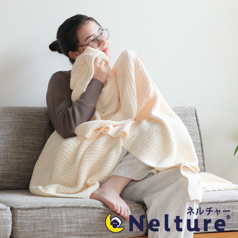 一位女性坐在沙發上，身著長袖上衣與長褲，蓋著一條奶油色、觸感柔軟、帶有條紋織紋的絨毛毯。