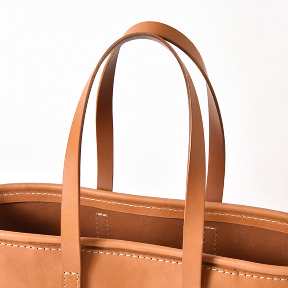 職人手作皮革經典托特包 T-9 L，採用天然棕色皮革製成，配有兩個長提把，包身邊緣及提把接縫處皆有白色縫線，展現細緻工藝。