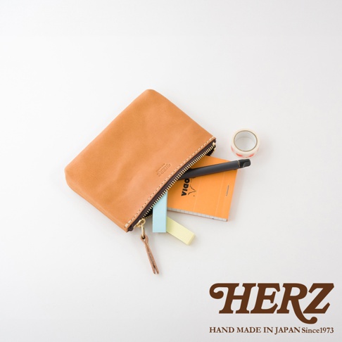 職人手作皮革長方形拉鍊收納包，材質為皮革，顏色為自然棕色，拉鍊為深色，包內裝有筆、筆記本和便利貼，旁邊有一個小卷筒膠帶。