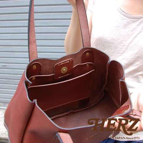 一件紅棕色皮革手提包，內部有兩個隔層和一個磁扣，由一個人拿著，背景是金屬捲門。