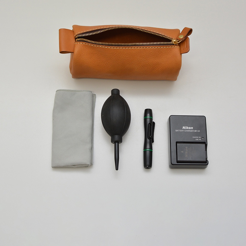 一個棕色皮革製成的拉鍊筆袋，旁邊擺放著一個灰色拭鏡布、一個黑色橡膠吹氣球、一支黑色綠色飾條的清潔筆，以及一個黑色 Nikon 電池充電器 MH-24，內含一顆 Nikon 電池。