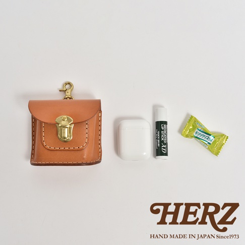 職人手作皮革耳機收納包 KF-4，淺棕色皮革，附黃銅鎖扣及掛鉤，旁為白色無線耳機、藥用護唇膏及綠色糖果。