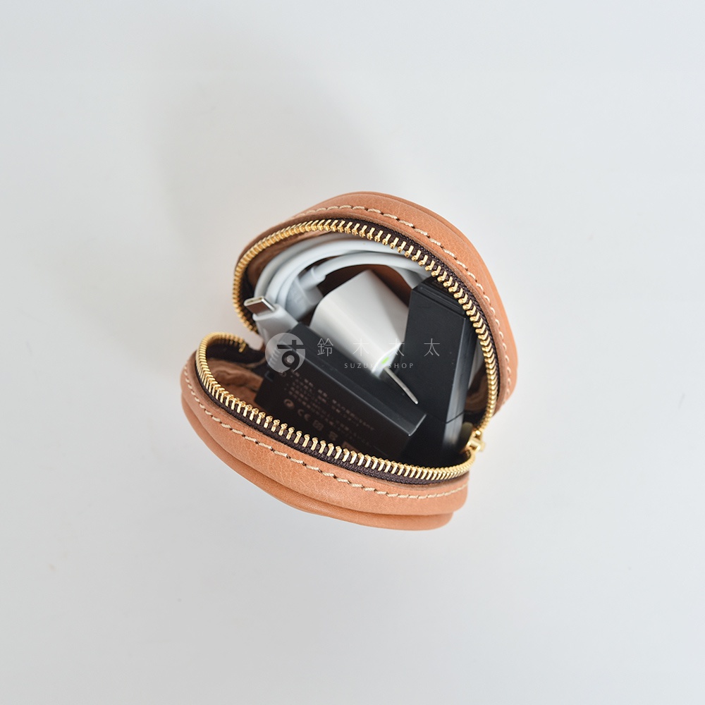 職人手作的皮革小物收納包，呈淺棕色，圓潤的三角飯糰造型，縫線細緻，內裝有充電器、充電線等電子配件，搭配金色拉鍊。