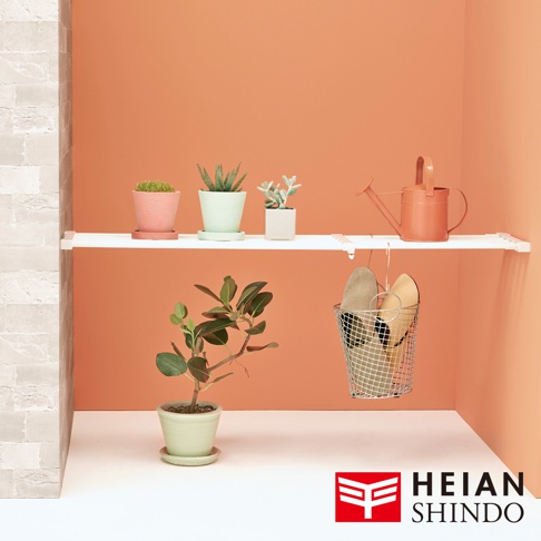 白色層架上擺放著粉色、淺綠色和白色盆栽植物，以及一個橘色澆水壺。層架下方懸掛著一個金屬網籃，籃內有兩雙鞋子。背景為橘色牆面。