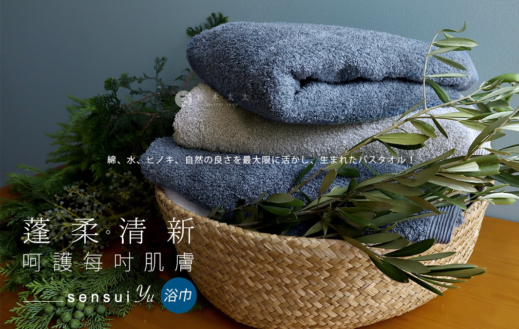 堆疊的藍色與灰色精梳棉浴巾，部分被橄欖枝和綠葉覆蓋，置於編織籃中。