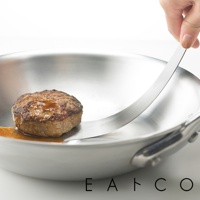 日製金屬食物鏟，鏟面上放著一個煎好的漢堡肉排，肉排表面有醬汁，鏟子伸入鍋中，鍋身為不鏽鋼材質。
